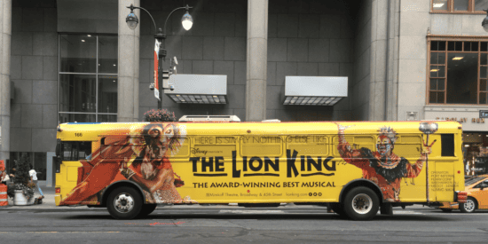 Grande ônibus amarelo fotografado na cidade com sinalização de promoção do Musical Rei Leão exibida no ônibus