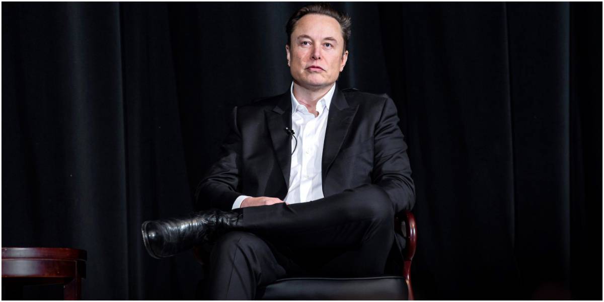 Elon Musk sentado de terno em frente a um fundo preto